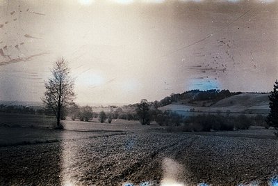 Vintage sepia-toned landscape showing open fields, sparse trees, and rolling hills under hazy skies. Likely early 20th-centur...