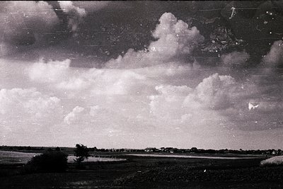Vintage black-and-white landscape featuring dramatic cumulus clouds over flat terrain. Low-lying fields and sparse trees sugg...