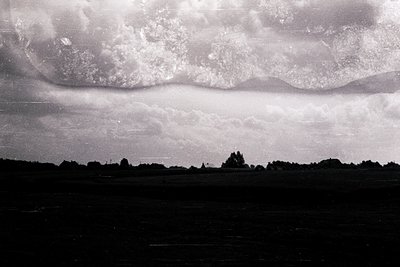 Vintage black-and-white landscape featuring dramatic low-hanging clouds over an open, flat field. Distant tree line and silho...