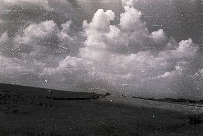Vintage black-and-white aerial view of open terrain with scattered trees and rolling hills under dramatic cumulus clouds. Fil...