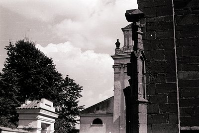 Black-and-white shot of a weathered stone bell tower with ornate Gothic-style detailing, including a cross-topped spire and d...