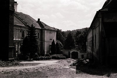 A decaying courtyard of a historic European estate, likely mid-20th century. Two-story buildings with peeling plaster, arched...