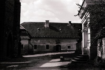 A weathered courtyard scene featuring a two-story building with a pitched roof and arched doorway. The structure’s plaster sh...