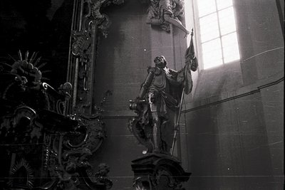 Baroque-style statue of a robed figure holding a raised sword, illuminated by natural light through arched windows. Intricate...