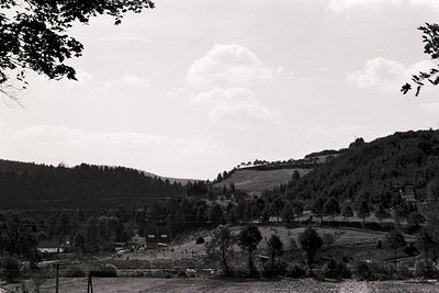Mid-20th century rural landscape with rolling hills, dense forest, and scattered farmland. Low-angle shot captures sparse str...