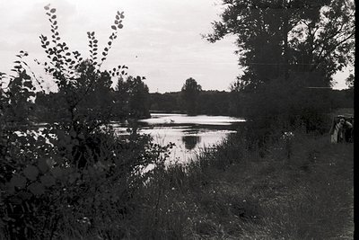Black-and-white lakeside scene with dense foliage framing the foreground. A small boat floats on calm water, surrounded by re...