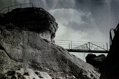 Black-and-white shot of a suspension bridge spanning rugged cliffs, framed by dramatic rock formations. The bridge’s cables a...