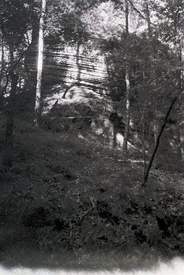 Vintage sepia-toned photo of a rural forest clearing with a lone figure in mid-stride, likely mid-20th century. The person we...