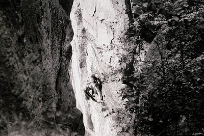 Black-and-white close-up of a rock climber ascending a sheer limestone face, mid-move with chalk dust visible. The texture of...