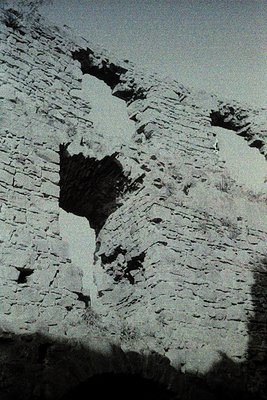 Rough-hewn stone wall with a narrow, vertical crack revealing dark interior. Likely historic masonry, possibly or earlier. Te...