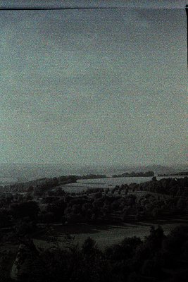 Vintage black-and-white landscape showing rolling farmland with scattered trees and a distant horizon. Low-contrast sepia ton...