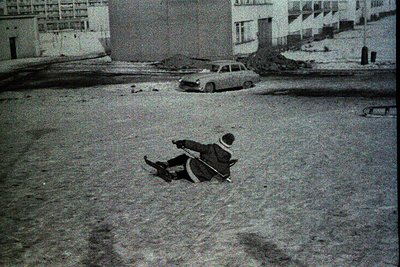Child sledding on snow-covered pavement, mid-1960s urban setting. Surrounding mid-rise concrete buildings and parked vintage ...