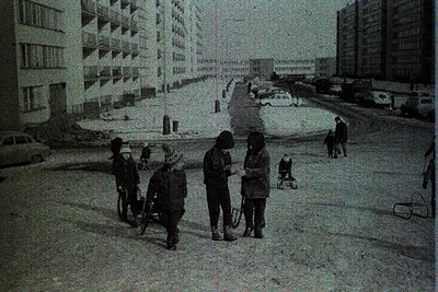 Mid-20th century urban street scene featuring Soviet-era concrete apartment blocks. Four children push a handcart laden with ...