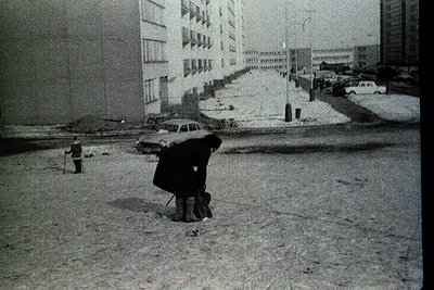 Snow-covered urban street with mid-century concrete apartment blocks. A lone figure in winter attire kneels, shoveling snow n...