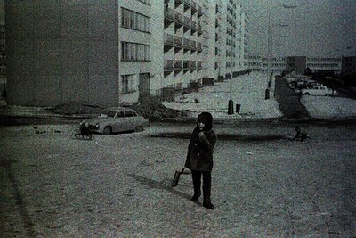 A child in winter attire stands on a snow-covered street, holding a shovel. Soviet-era apartment blocks and a parked car domi...
