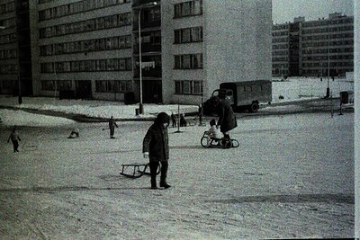 Black-and-white street scene featuring children sledding on snow-covered pavement, flanked by mid-rise apartment blocks. Snow...