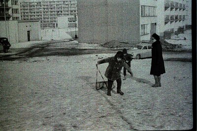 A black-and-white snapshot captures two children sledding on a snow-covered street in a Soviet-era urban setting. The child i...