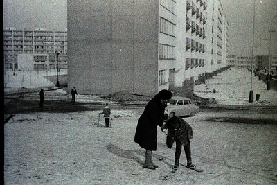 A woman in a long coat and headscarf assists a child with a backpack in a snowy urban setting, likely Eastern Europe, 1960s–1...
