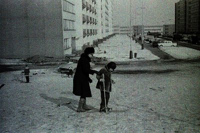 A woman and child in winter coats examine snow-covered pavement beside a Soviet-era apartment block. Snowbanks line the stree...