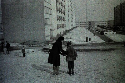 A woman and child in winter coats stand on a snow-covered street, holding hands. High-rise apartment blocks with shuttered wi...