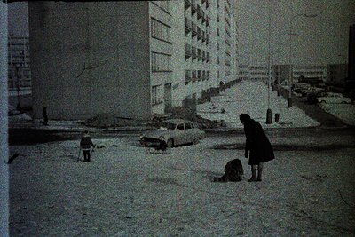 Snow-covered urban street with mid-rise concrete apartment blocks, likely Eastern Bloc architecture. Two individuals—one knee...