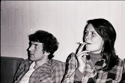 Black-and-white portrait of two individuals in a relaxed indoor setting, likely 1970s. The woman wears a plaid shirt and hold...