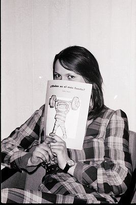 Black-and-white portrait of a young woman holding a children’s book titled *"¿Quién es el más fuerte?"* ("Who is the stronges...