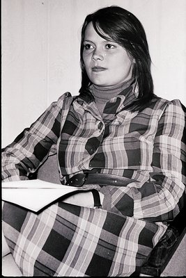 Portrait of a person in a plaid shirt (checkered pattern) and turtleneck, seated indoors with a book or notebook in lap. Mid-...