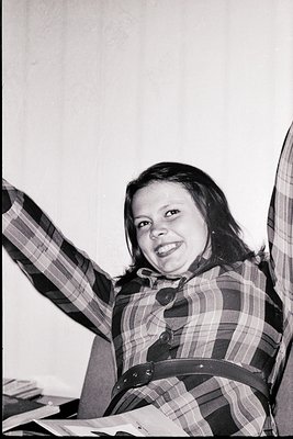 Vintage black-and-white portrait of a cheerful individual in a plaid shirt, seated indoors with one arm raised. The setting a...