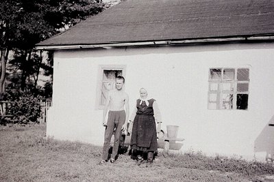 Two individuals pose outside a modest, single-story house with whitewashed walls and simple window frames, likely mid-20th ce...