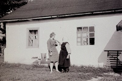 Two women in mid-20th-century attire stand beside a modest, whitewashed house with lattice-pane windows. One adjusts the othe...