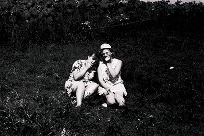 Two individuals pose intimately in a grassy outdoor setting, likely mid-20th century. The woman wears a floral-patterned dres...