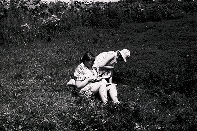 Two individuals embrace in a grassy field, mid-20th century. The woman wears a patterned dress and hat; the man, a light-colo...