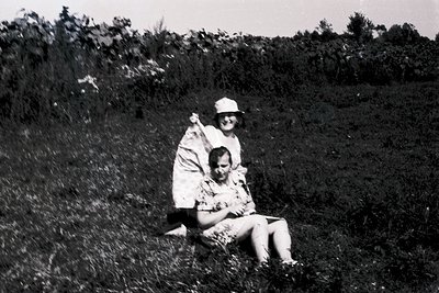 Black-and-white outdoor portrait of two individuals in a grassy field, likely mid-20th century. The adult wears a wide-brimme...