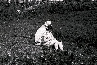 Mid-20th century black-and-white photo of two children sitting close in grassy field. Child on left wears patterned dress and...