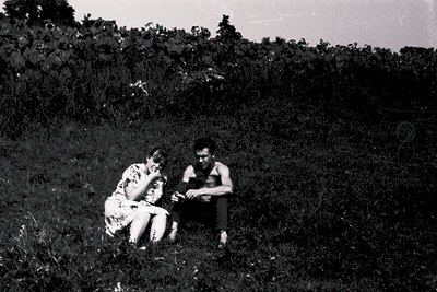 Black-and-white candid of two individuals seated on grassy hillside, mid-20th century. Woman in floral dress, man in sleevele...
