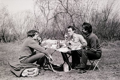 Three men seated around a folding camp table in a wooded area, sharing a meal in the 1960s–1970s. Mid-century camping gear—fo...
