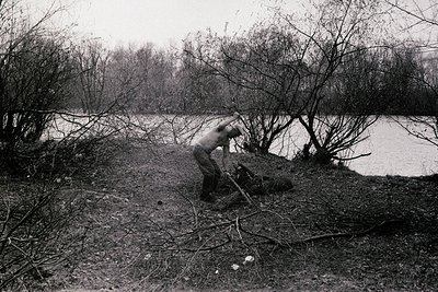 Black-and-white photograph of a lone figure clearing fallen branches from a riverside path, likely post-industrial or rural. ...