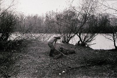 Black-and-white image of a lone individual clearing fallen branches from a riverside path, surrounded by bare trees and muddy...