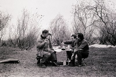 Three individuals gather around a small campfire table in a leafless forest, mid-conversation. Mid-century outdoor attire sug...
