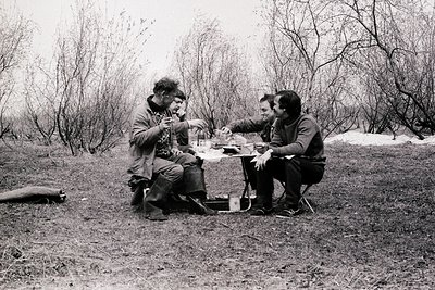 Two men share a meal outdoors around a small campfire, seated on a rock. One holds a mug, the other a plate with food. Mid-20...