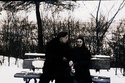 Couple seated on a wooden bench in a snowy park, mid-20th century. Man in dark coat, woman in long dark coat and headscarf. S...