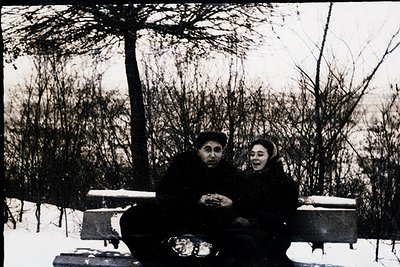 Vintage black-and-white photo of two individuals seated on a snow-covered bench in a wooded area, likely mid-20th century. Th...