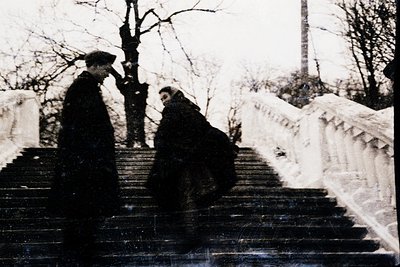 Two individuals converse on snow-covered stone steps in a park-like setting, mid-20th century. The man on left wears a cap an...