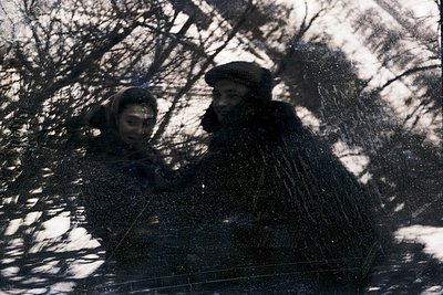 Silhouetted figures in heavy winter coats and hats, partially obscured by bare trees, suggest rural winter activity. The mono...