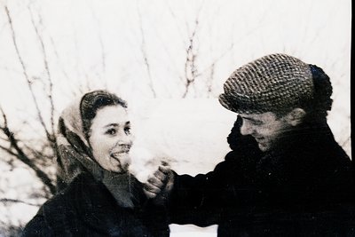 Mid-20th century black-and-white photo of a man and woman in winter attire—her in a headscarf, him in a flat cap—posing outdo...