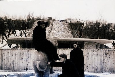 Vintage black-and-white photo of a man atop a polar bear in a snowy enclosure, assisted by another man in mid-reach. A woman ...