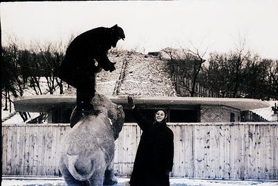Mid-20th century black-and-white photo of a circus act: a man balances atop a seated elephant, while a woman in a long coat p...