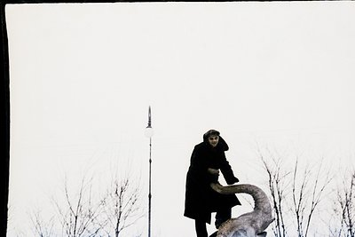 Black-and-white portrait of a person in a long coat leaning against a stylized, abstract sculpture resembling a reclining fig...