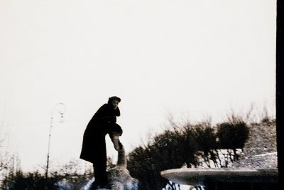 Black-and-white street scene featuring a lone figure in a long coat and cap bending to tie a boot in an urban setting. Mid-ce...